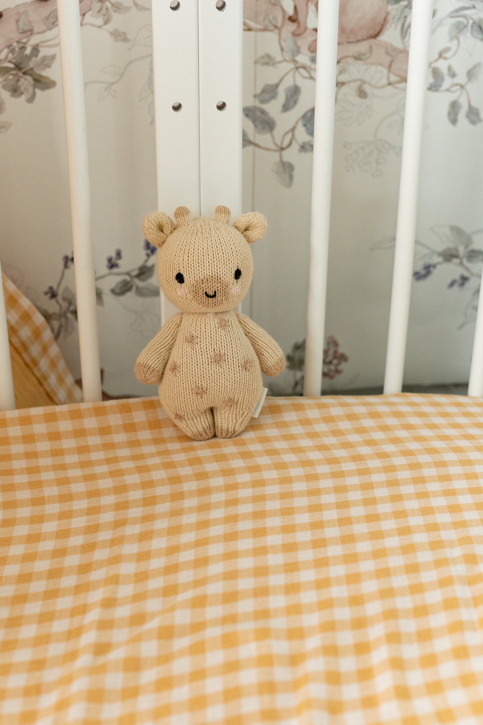 Knitted teddy bear on a yellow checkered blanket in a crib with floral wallpaper.
